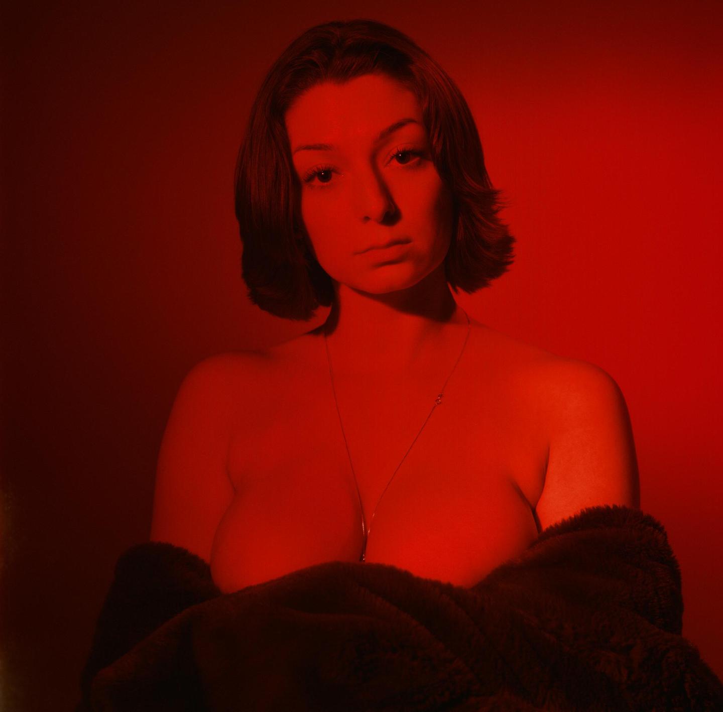 A woman with short hair and a necklace is illuminated by red light, wearing a dark, furry garment.