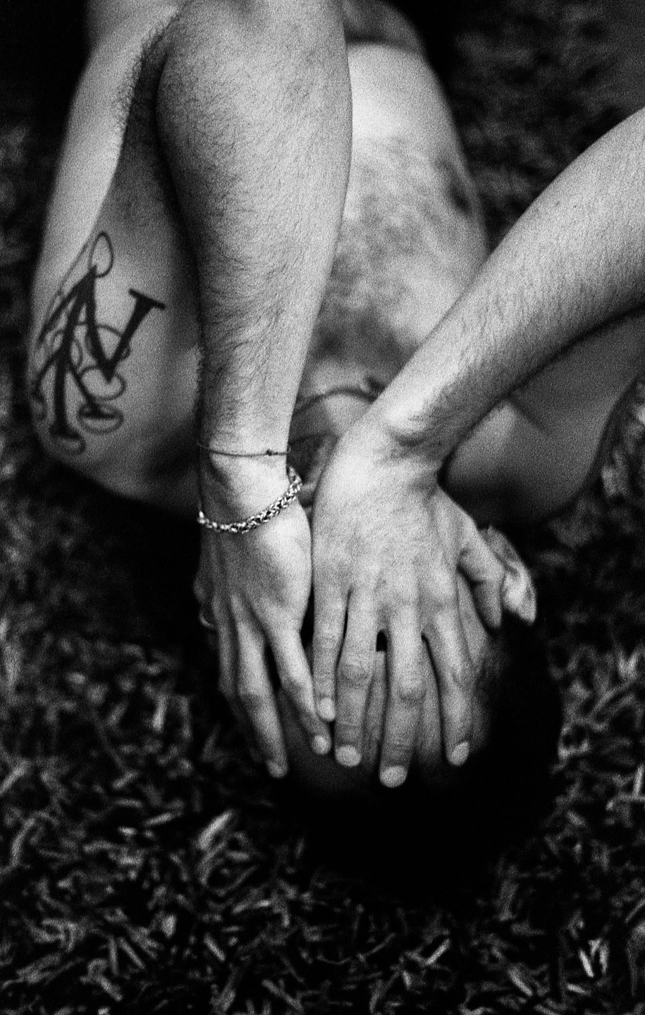 A man lies on the ground with his hands covering his face, revealing a tattoo on his shoulder and a bracelet on his wrist.