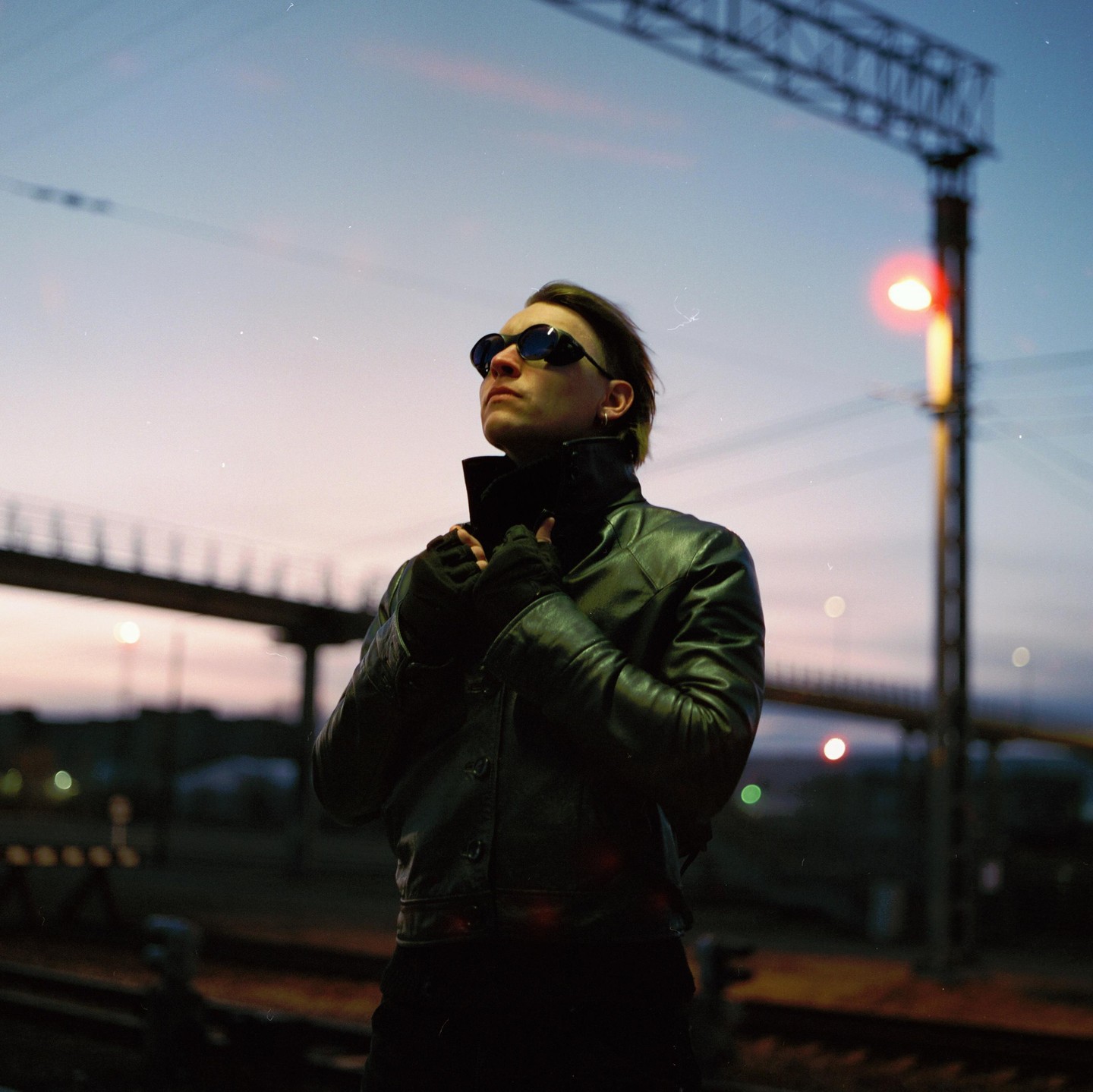 A person in a leather jacket and sunglasses stands on a railway platform at dusk.