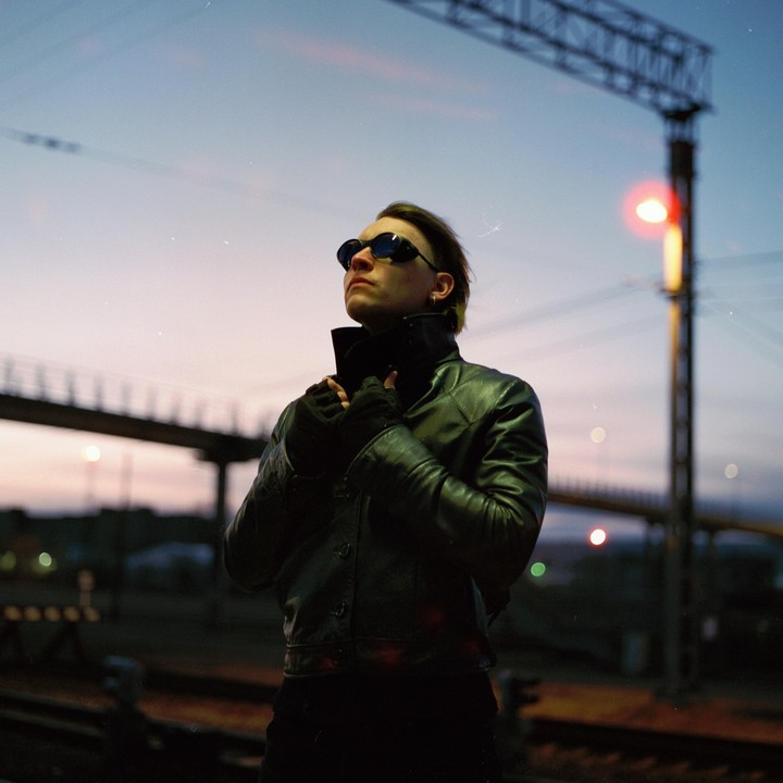 A person in a leather jacket and sunglasses stands on a railway platform at dusk.