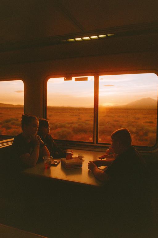 Three people sit at a table inside a train car as the sun sets over a vast landscape outside the window.