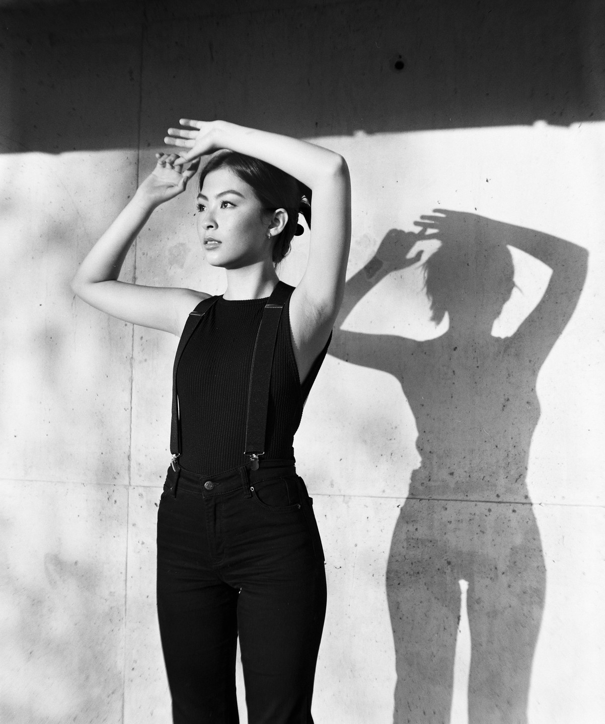 A woman in a tank top and suspenders poses against a concrete wall with her shadow cast beside her.