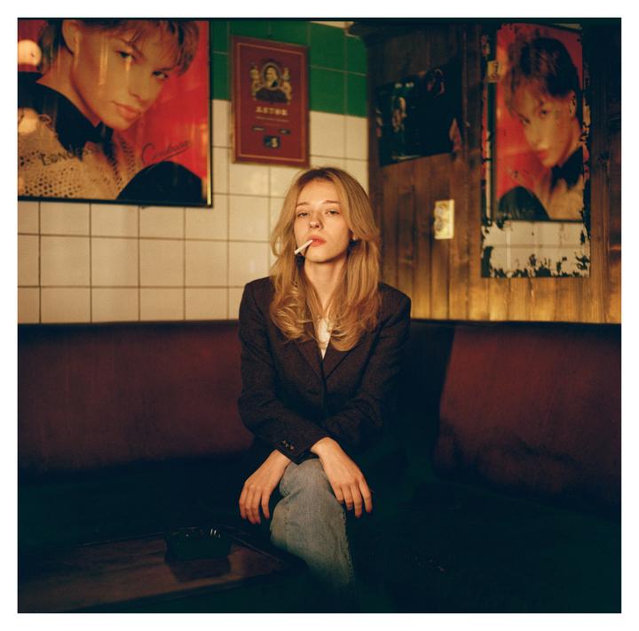 A woman with a cigarette in her mouth sits on a dark red couch in a dimly lit room with posters on the wall.