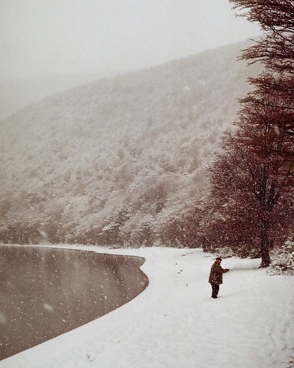 A man stands on a snow-covered shore next to a lake with a snowy forest and mountain in the background.