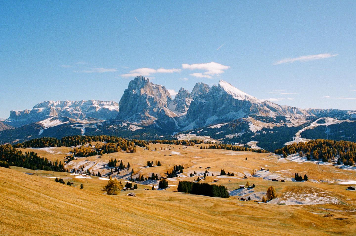 A vast landscape shows snow-capped mountains towering over a valley with patches of snow and autumn trees.