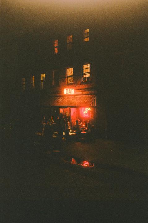 A group of people are gathered outside a bar with a neon sign at night.