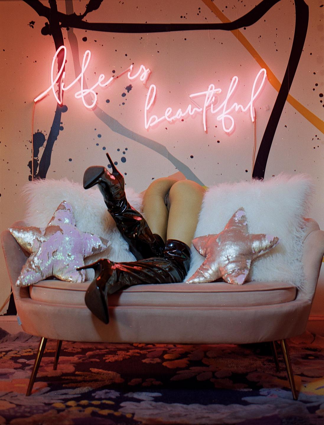 A person wearing shiny black boots is lying on a pink couch with a neon sign that reads "life is beautiful" on the wall.