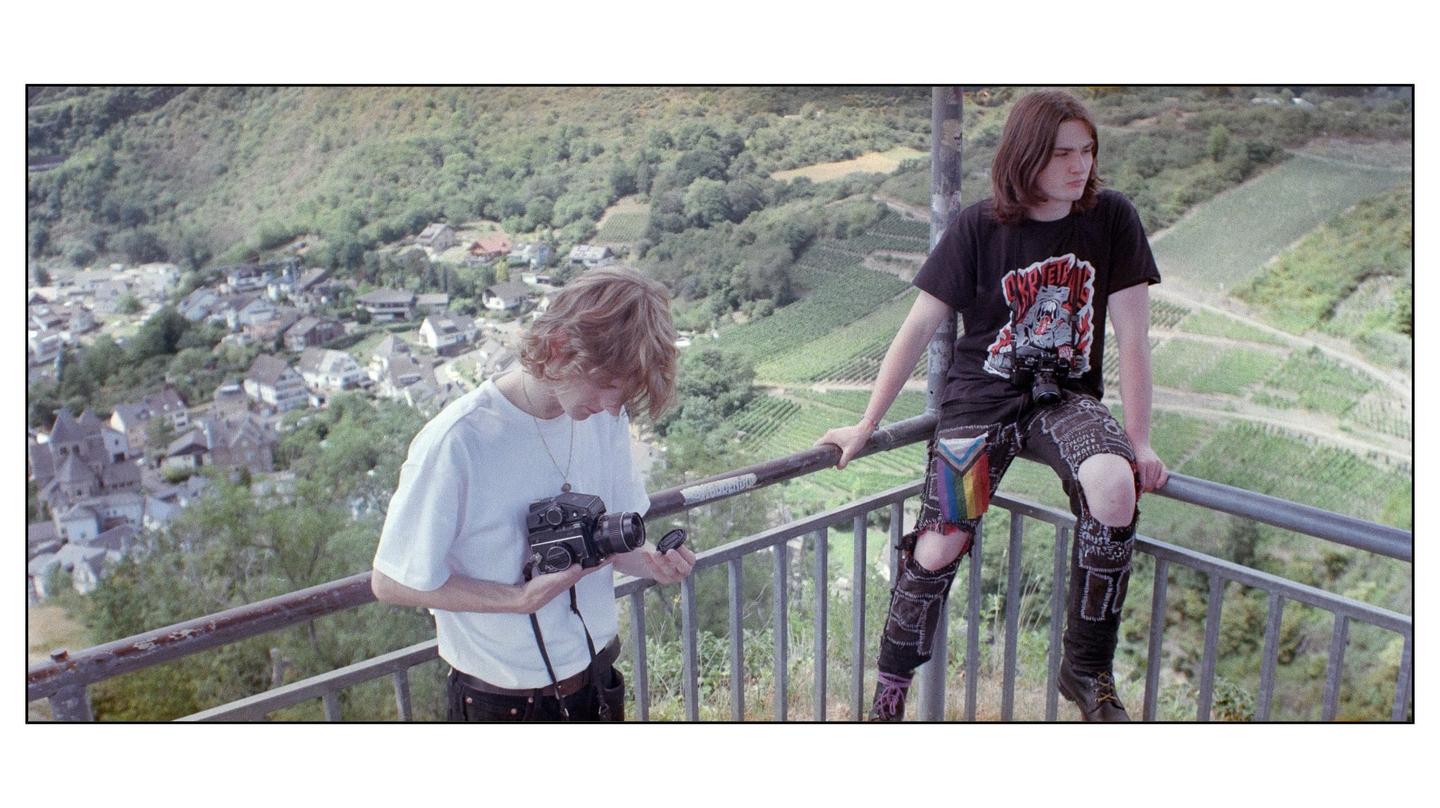 Two men are on a balcony overlooking a village and vineyards.