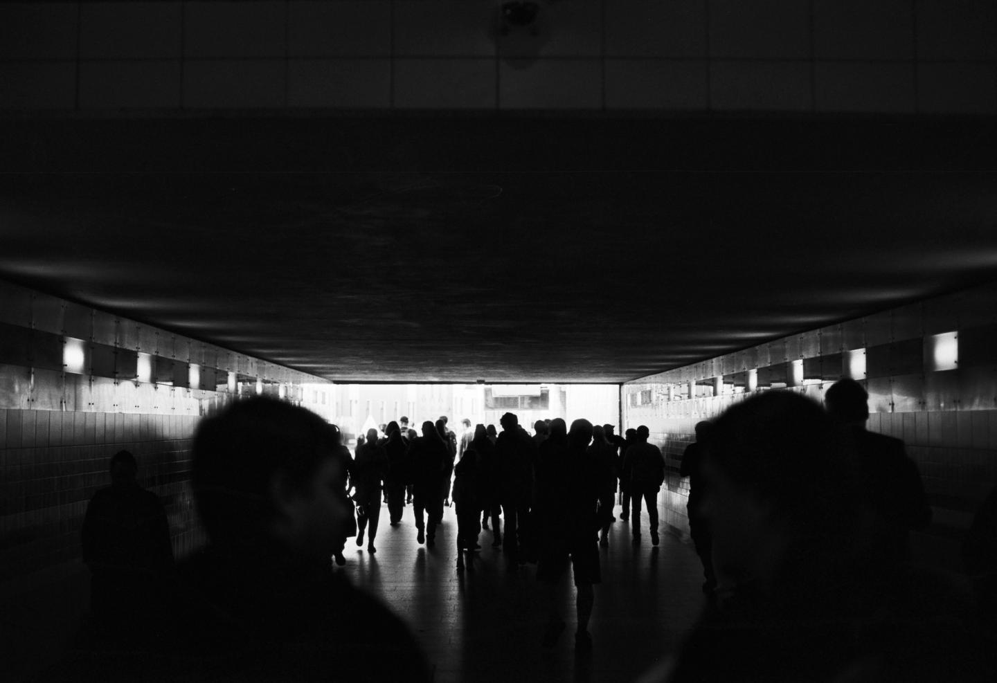 A crowd of people walks through a dark tunnel towards a bright light at the end.