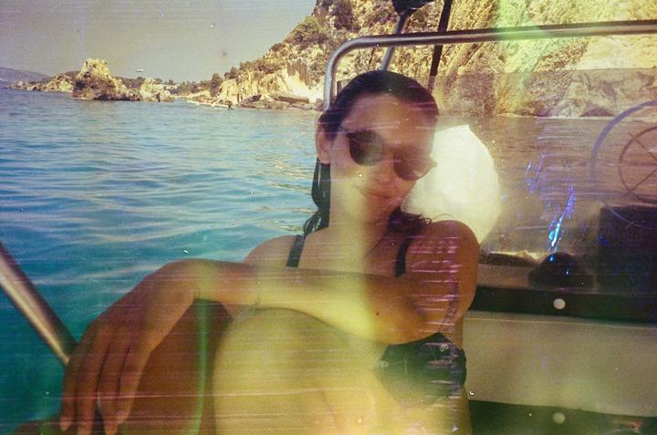 A woman wearing sunglasses sits in a boat on the water with a rocky coastline in the background.