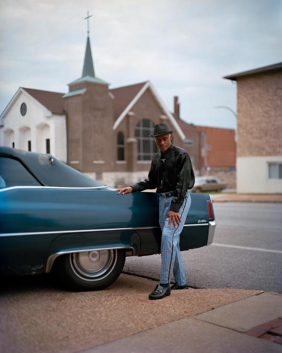 A man in a hat and plaid pants stands next to a blue car on a street with a church in the background.