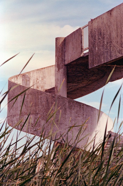 A concrete spiral staircase rises from behind tall grass against a blue sky.