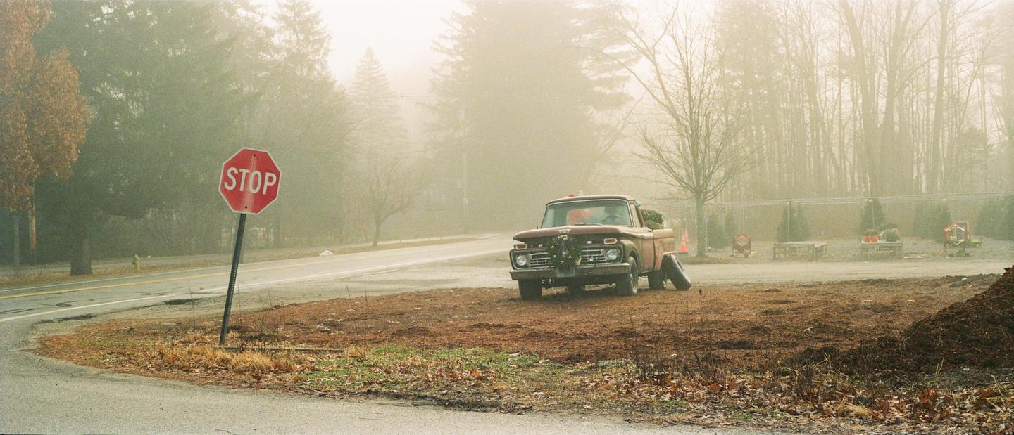 A red stop sign stands next to a road with a vintage truck parked on a dirt patch, all shrouded in fog.