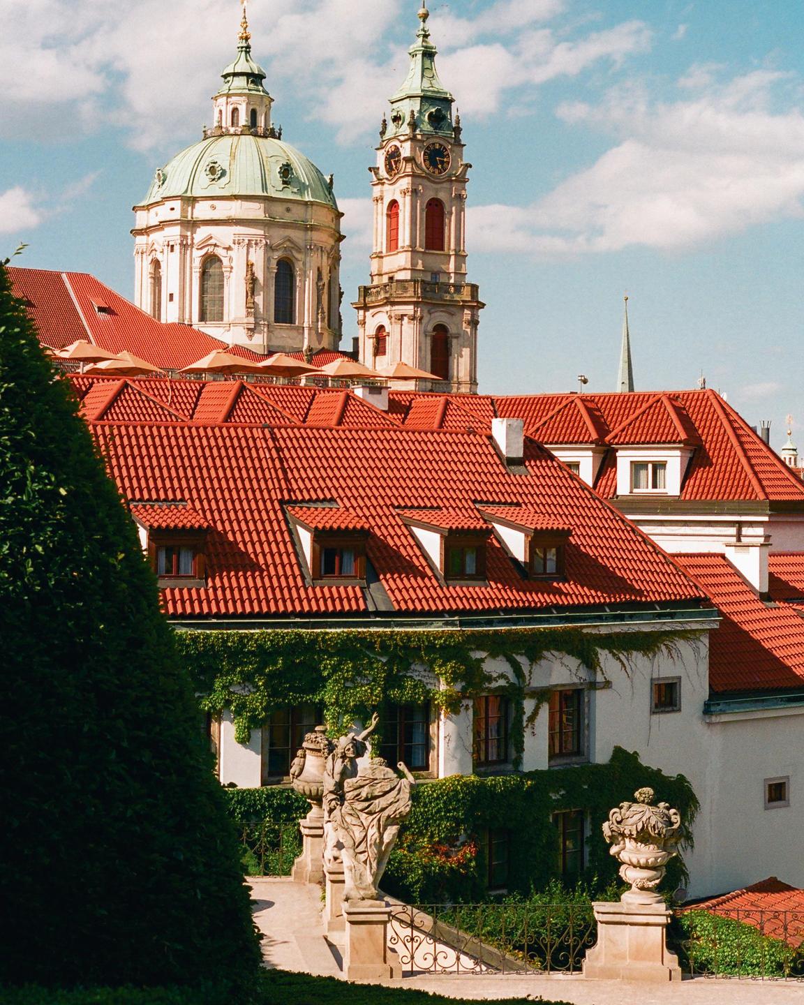 A view of red-tiled rooftops and a building covered in ivy, with a domed church and clock tower in the background.