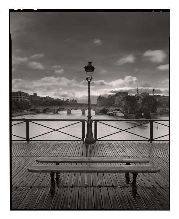A lamppost and two benches are on a wooden deck overlooking a river with bridges and buildings in the distance under a cloudy sky.