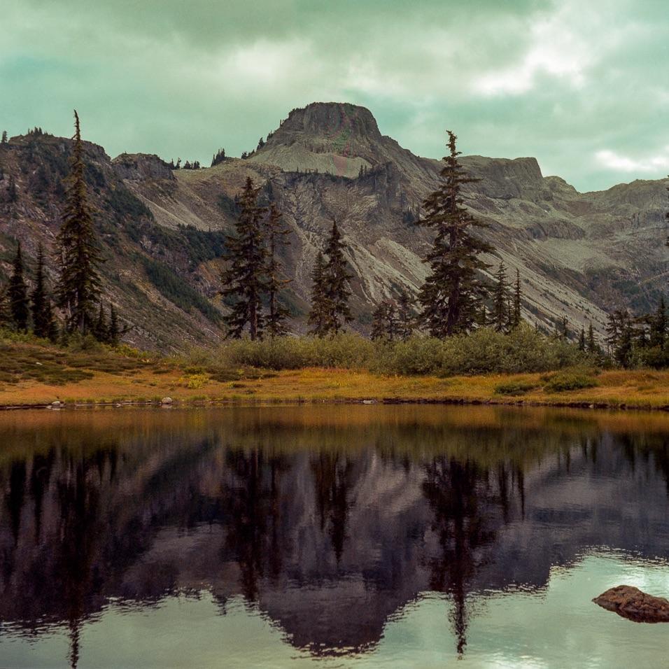 A mountain and evergreen trees are reflected in a still lake under a cloudy sky.