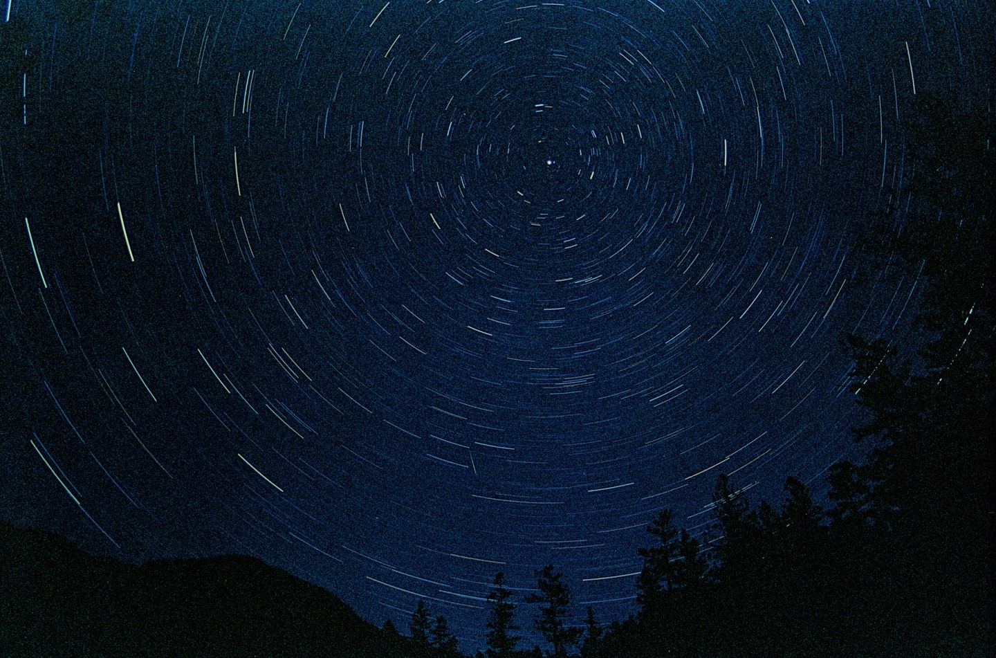 Star trails arc across the night sky above a dark mountain and forest.