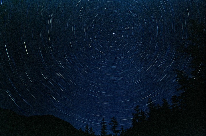 Star trails arc across the night sky above a dark mountain and forest.
