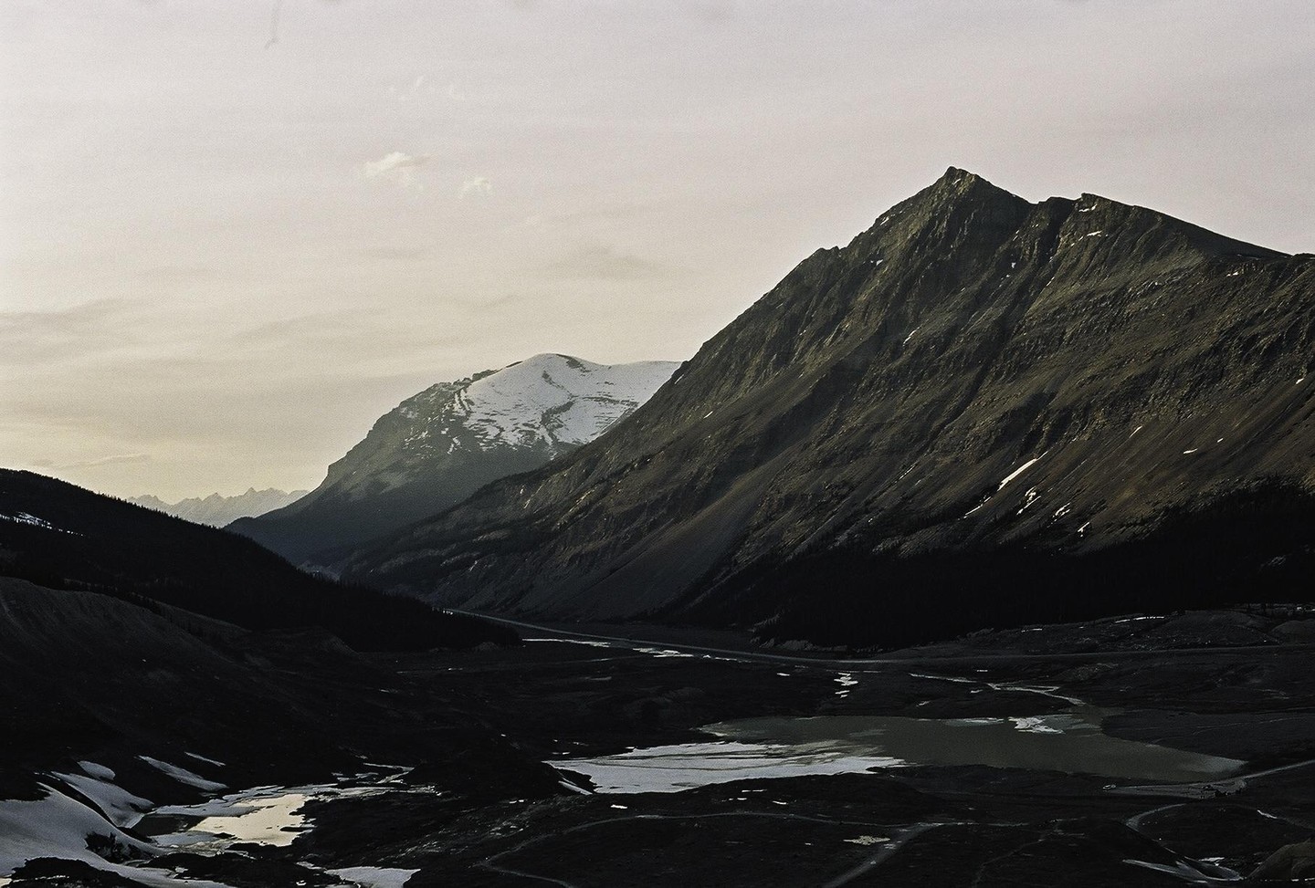 A valley with a road and a lake is nestled between two large mountains, one of which is snow-capped.