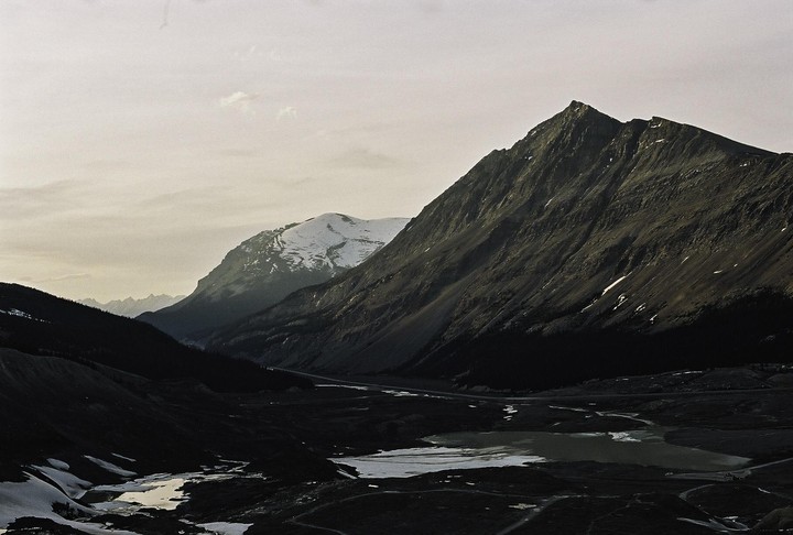 A valley with a road and a lake is nestled between two large mountains, one of which is snow-capped.