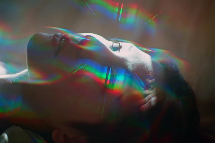 A woman lies with her head tilted back, her face illuminated by colorful light streaks.