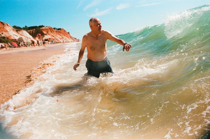 A man stands in the ocean as a wave breaks over him, with a sandy beach and cliffs in the background.
