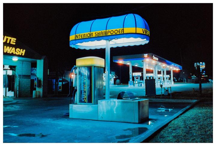 A blue and yellow awning for an interior shampooer stands in front of a gas station and car wash at night.
