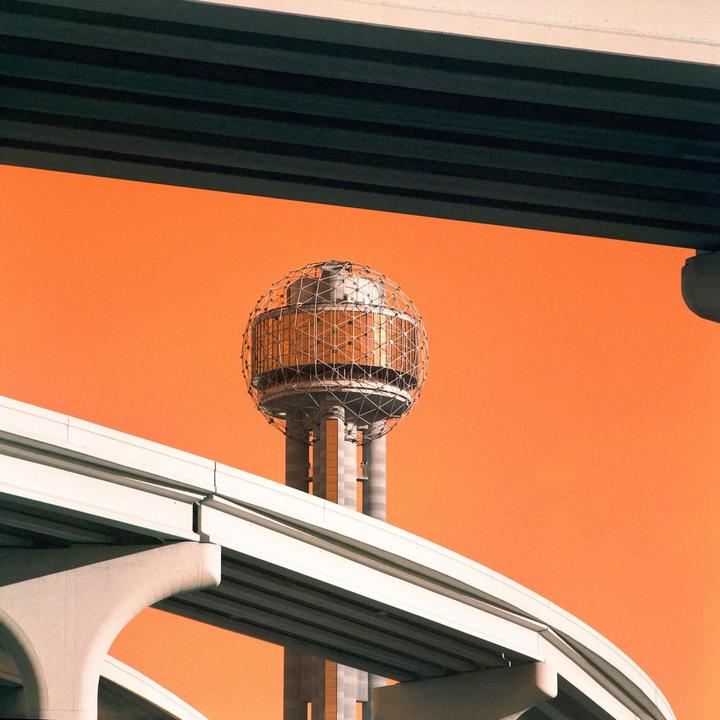 A tall tower with a spherical top is framed by two overpasses against an orange sky.