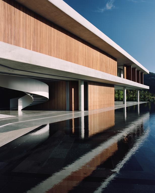 A modern building with wood paneling and concrete accents is reflected in a dark pool of water under a clear sky.