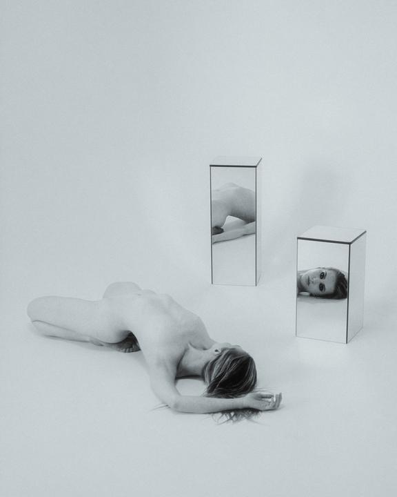 A woman lies on a white surface with two mirrored pillars reflecting her body and face.