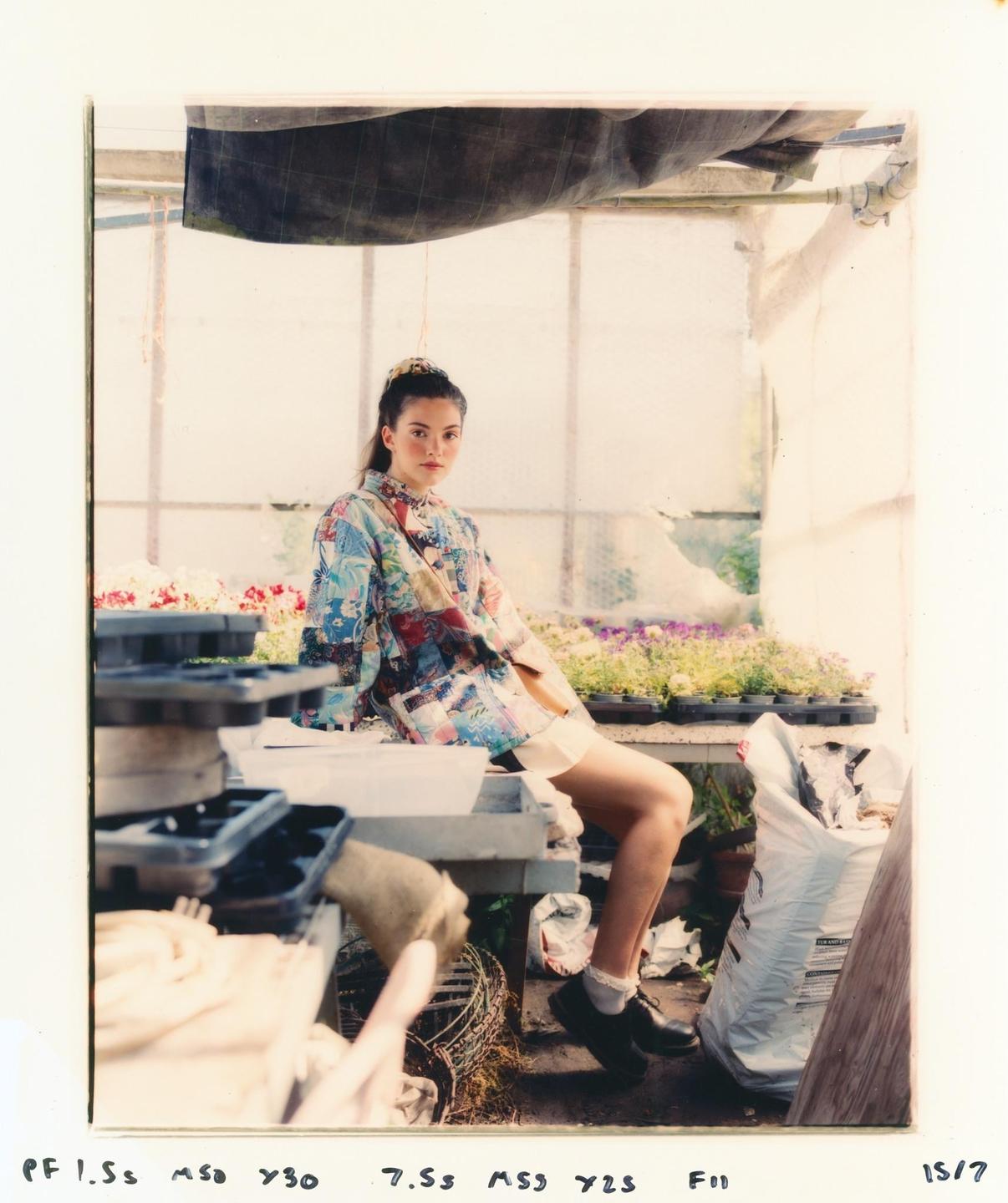 A woman wearing a colorful shirt sits on a table in a greenhouse with flowers and gardening supplies.