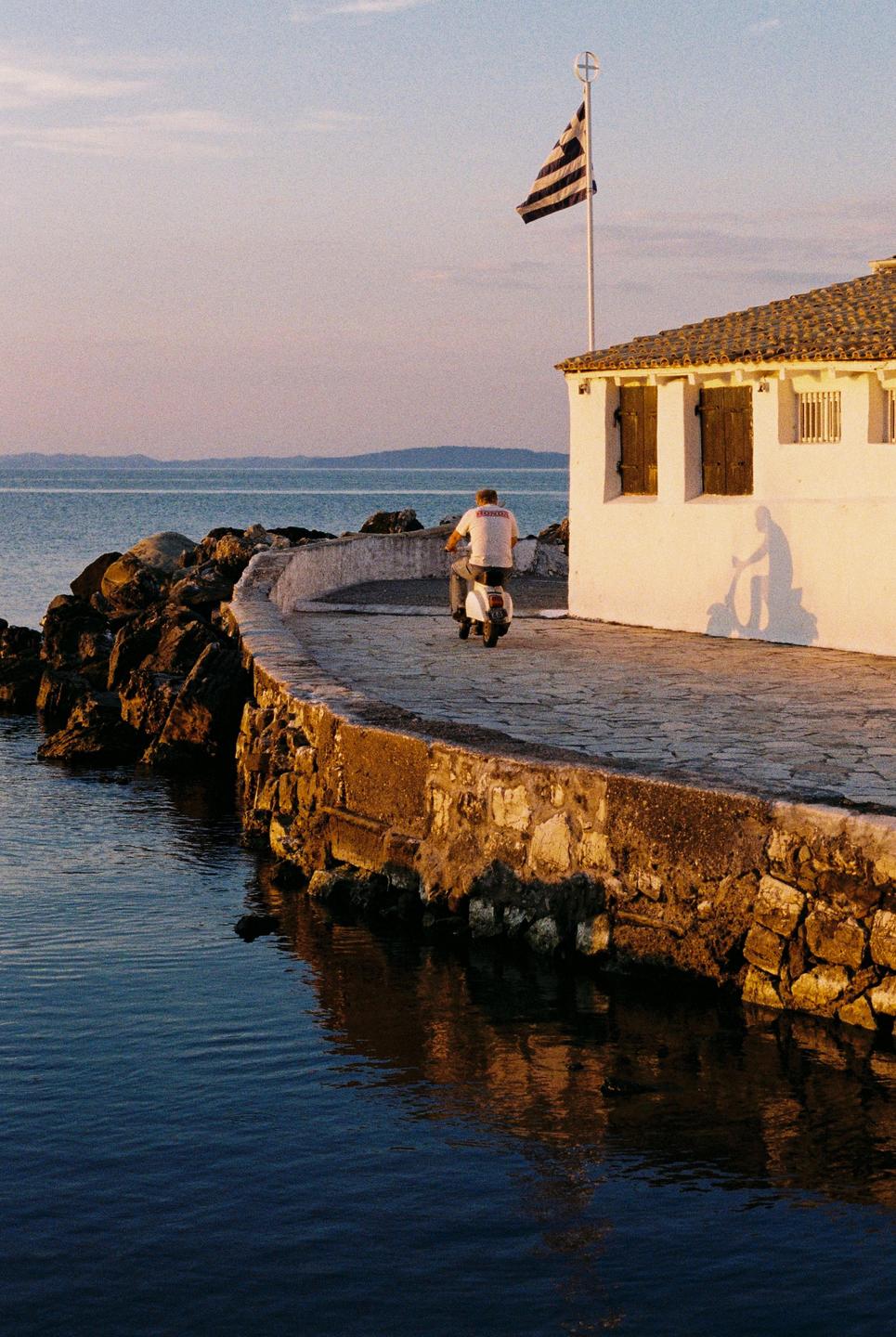 A man rides a scooter along a stone path next to a body of water with a white building and a flag.