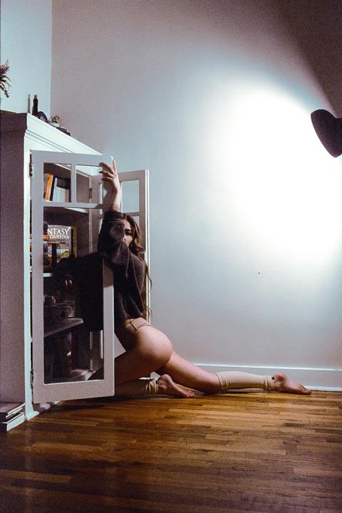 A woman in a sweater and underwear poses on a wooden floor next to a white bookshelf.