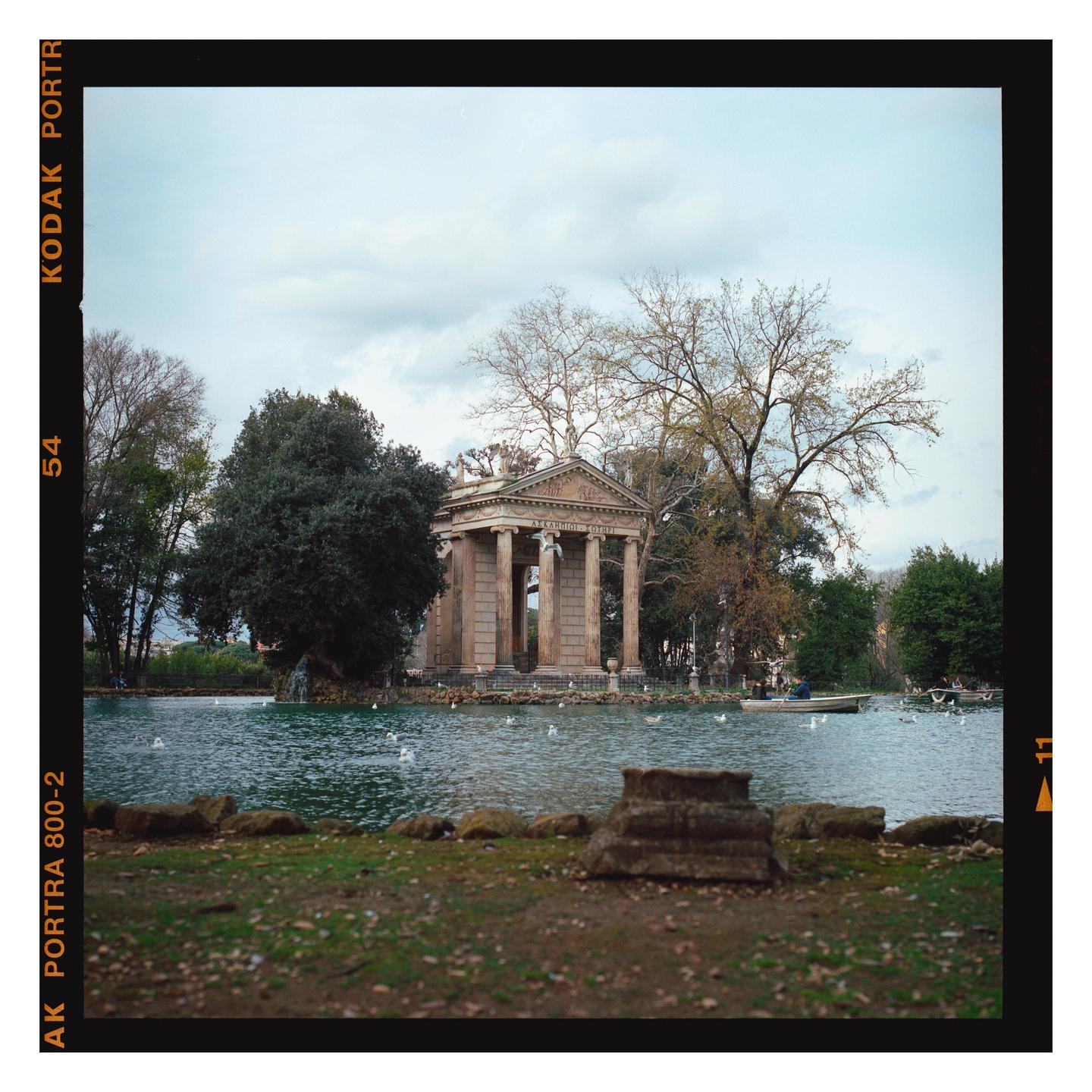 A classical temple with columns stands on an island in a lake, surrounded by trees and with a small boat on the water.