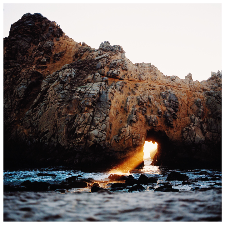 Sunlight streams through a natural arch in a large rock formation on a beach with waves crashing on the shore.