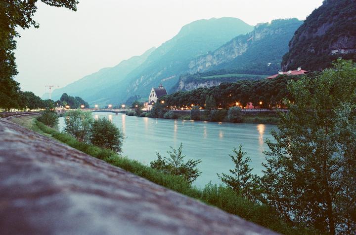 A river flows through a valley with mountains in the background and buildings and trees on its banks.