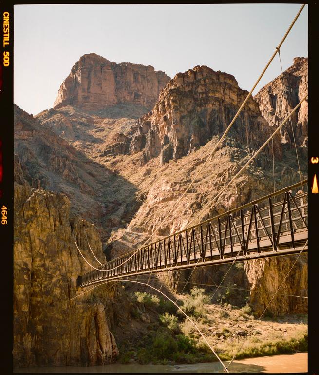 A suspension bridge spans a canyon with a river at its base, surrounded by towering rock formations under a clear sky.