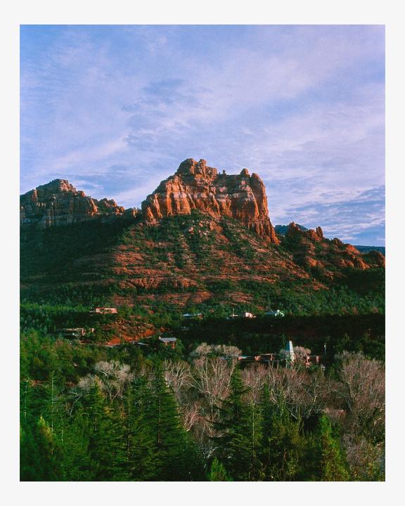 A large red rock formation rises above a forest with some buildings nestled among the trees.