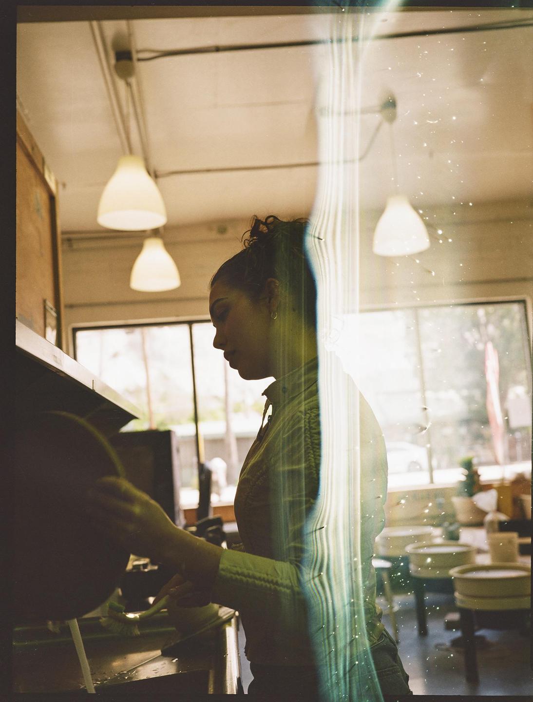 A woman with her hair in a bun washes dishes in a kitchen with hanging lamps and large windows.