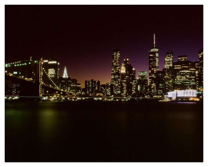 The New York City skyline and Brooklyn Bridge are illuminated at night, with reflections visible on the water.
