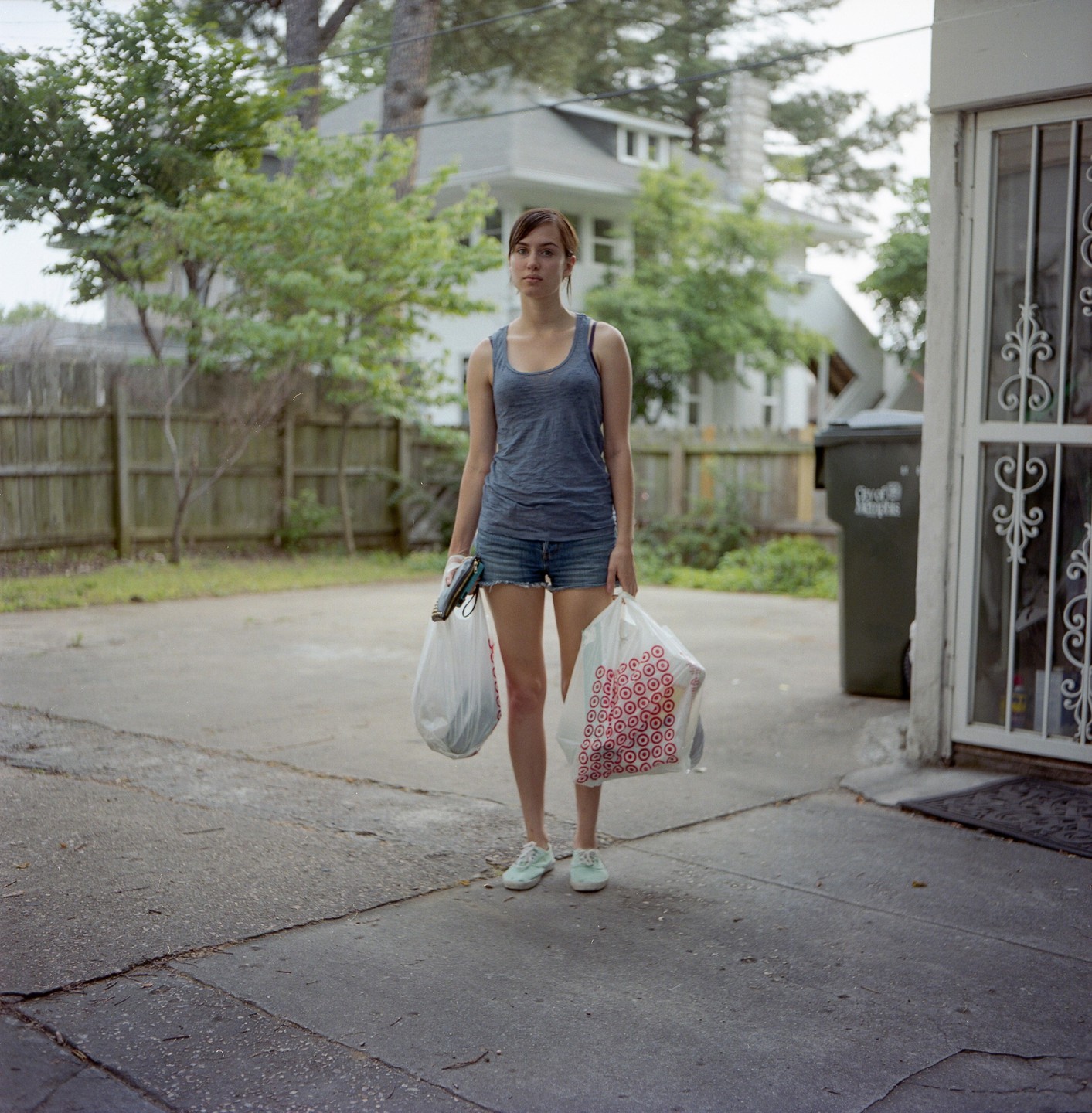 A woman stands in a driveway holding two shopping bags, with a house and fence in the background.