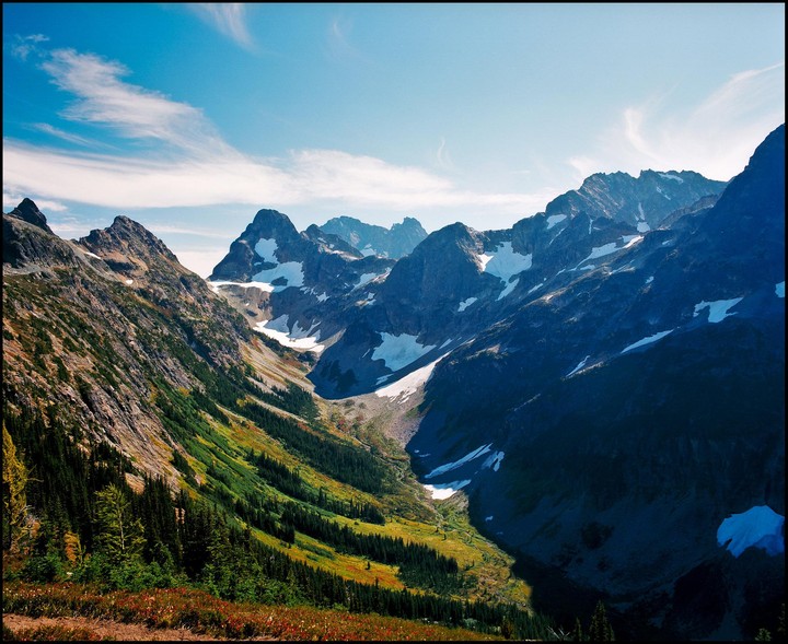 A valley with snow-capped mountains and evergreen trees is visible under a blue sky with wispy clouds.