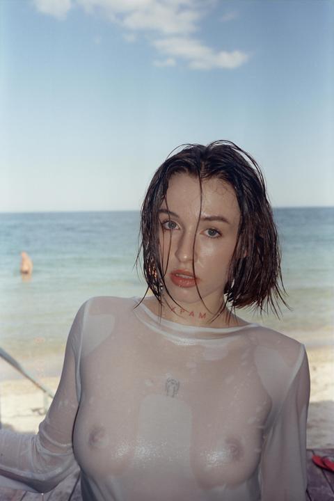 A woman with wet hair and a wet white shirt stands on a beach with the ocean and sky in the background.