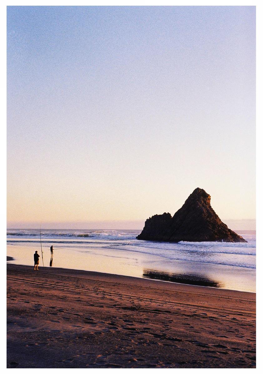 Two people stand on a beach with a large rock formation in the ocean at sunset.