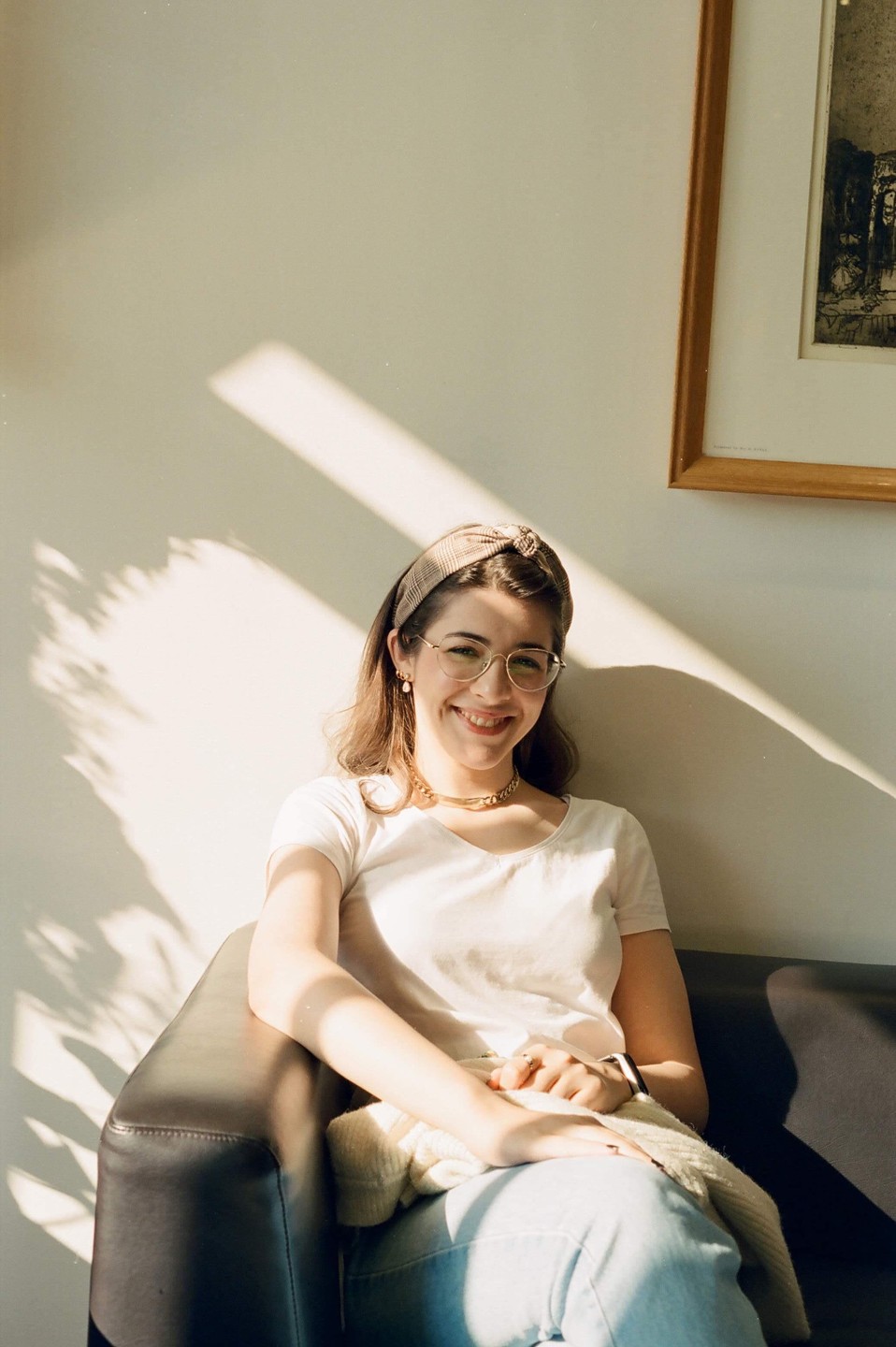 A young woman with a headband and glasses sits on a couch, smiling at the camera with sunlight and shadows on the wall behind her.