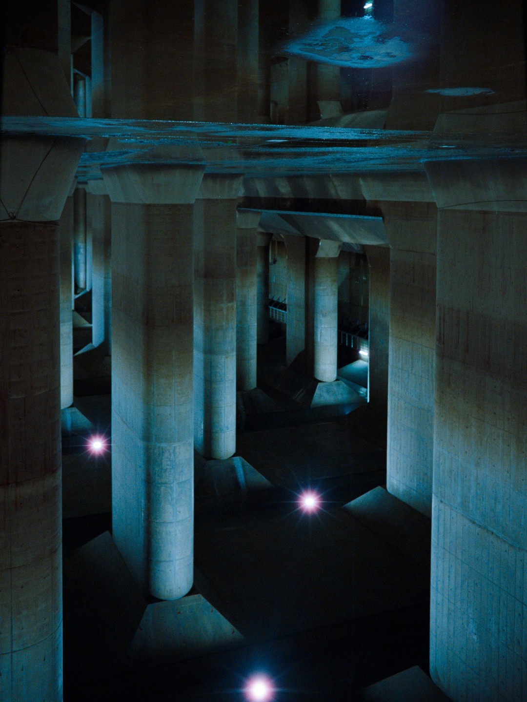 A vast underground structure with numerous concrete pillars is illuminated by blue light from above and several bright pink lights on the floor.