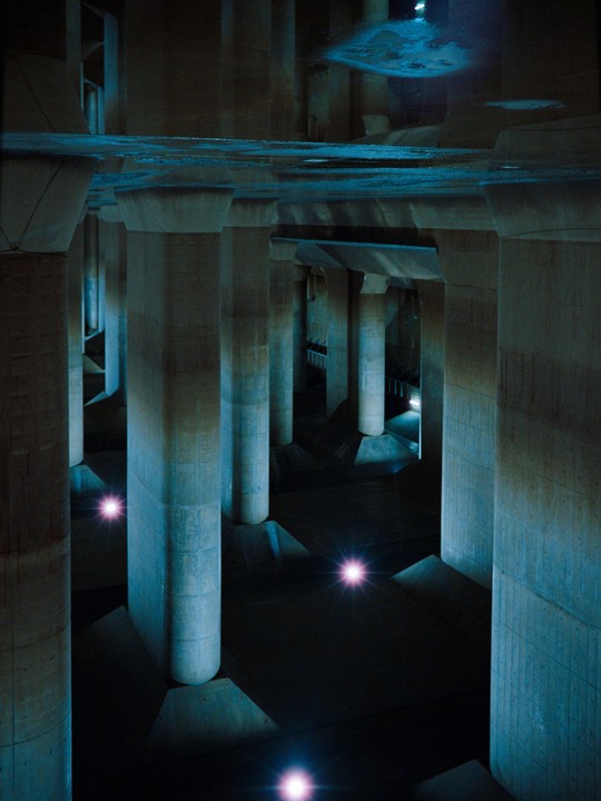 A vast underground structure with numerous concrete pillars is illuminated by blue light from above and several bright pink lights on the floor.