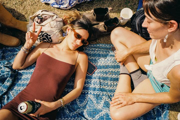 Two women are relaxing on a blue patterned blanket on the grass, one holding a can and making a peace sign.