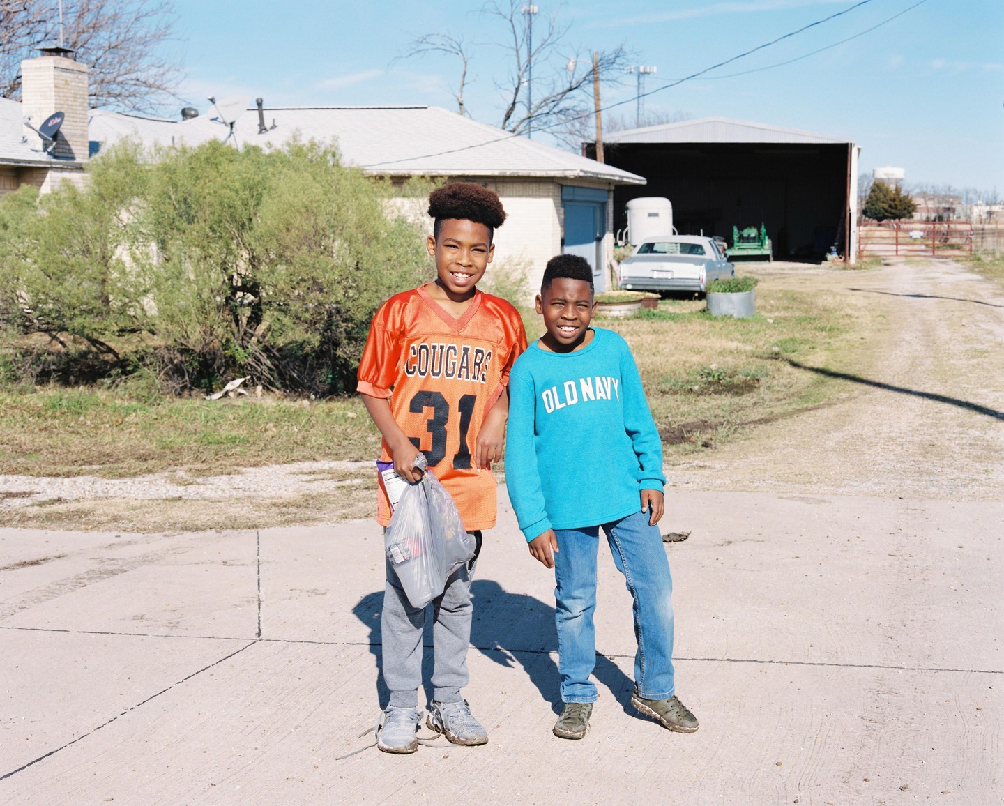 Two boys stand on a paved surface in front of a house and a shed.