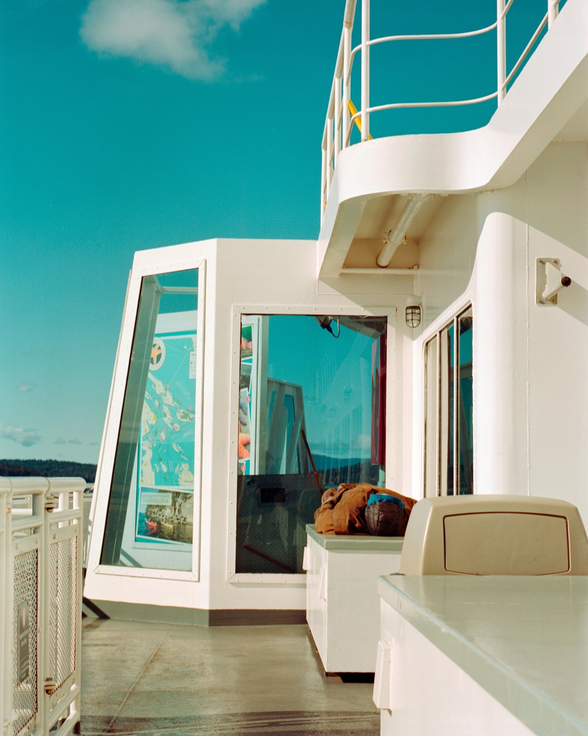 A brown bag rests on a white bench on the deck of a ferry under a bright blue sky.